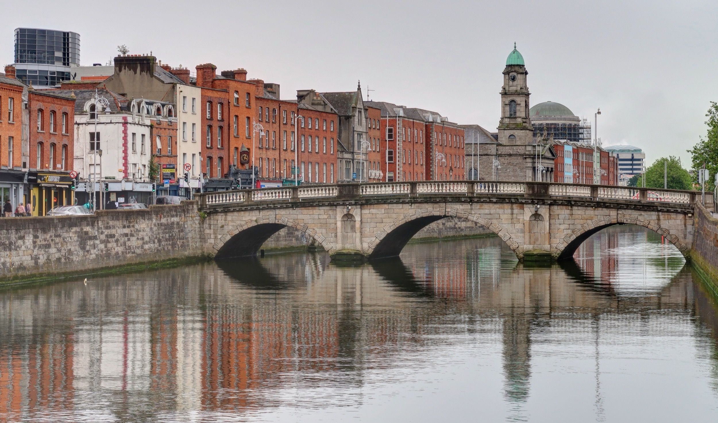 Blick über den River Liffey auf eine steinerne Bogenbrücke mit den charakteristischen roten Backsteingebäuden Dublins im Hintergrund.